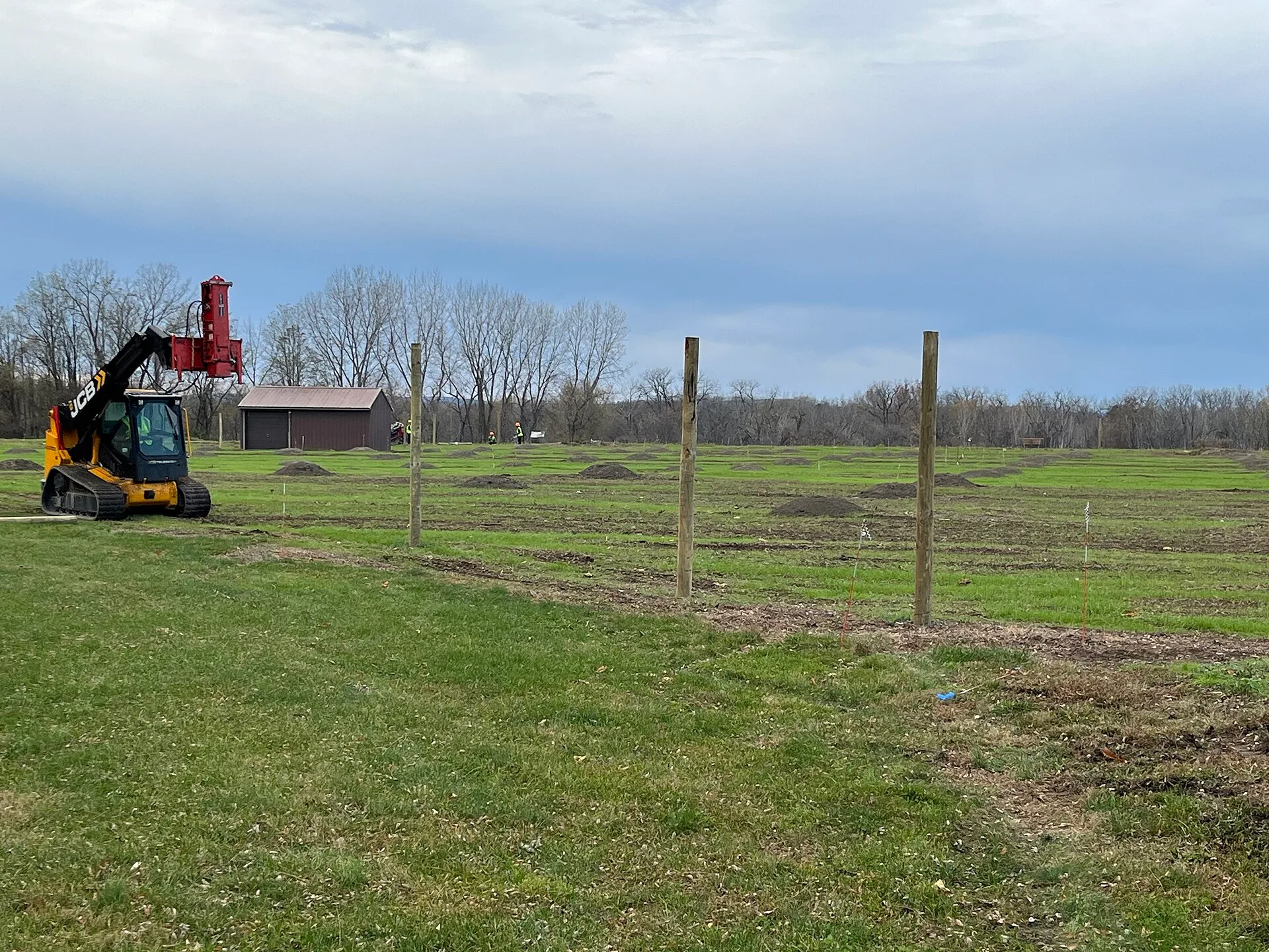 Tall wooden fence posts stand in front of the orchard area, with a fencing machine to the right. Installing the deer fencing around the orchard area. Post location was determined using GIS to avoid sensitive areas, and planners selected an option that would minimize visual intrusion. Keywords: orcha. Fence Contractor Customer Financing: Four Routes