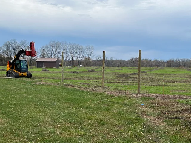 Tall wooden fence posts stand in front of the orchard area, with a fencing machine to the right. Installing the deer fencing around the orchard area. Post location was determined using GIS to avoid sensitive areas, and planners selected an option that would minimize visual intrusion. Keywords: orcha. Fence Contractor Customer Financing: Four Routes